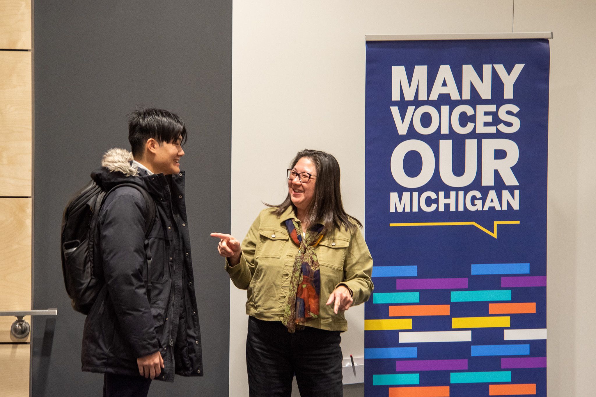 Two people laughing together in front of a colorful Many Voices Our Michigan banner