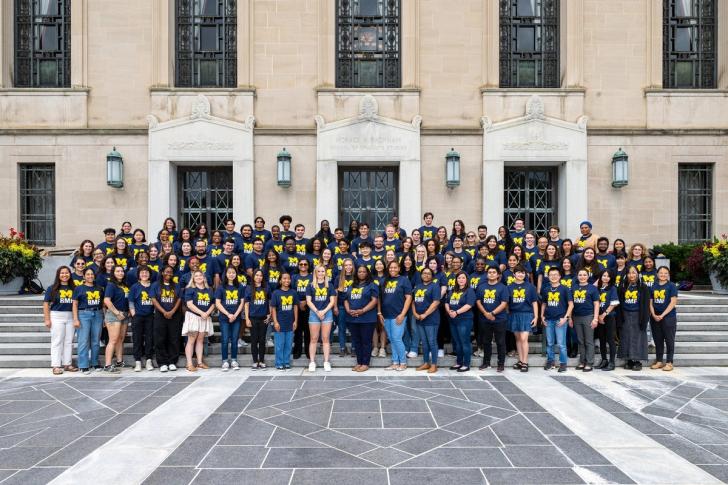 A group photo of people wearing Michigan shirts on the steps of the Rackham Building